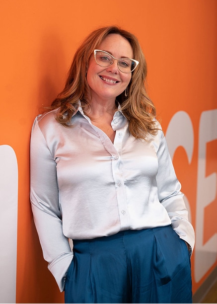 Katie smiling confidently in front of a bold orange wall, wearing a light satin blouse and blue trousers in a modern, professional portrait